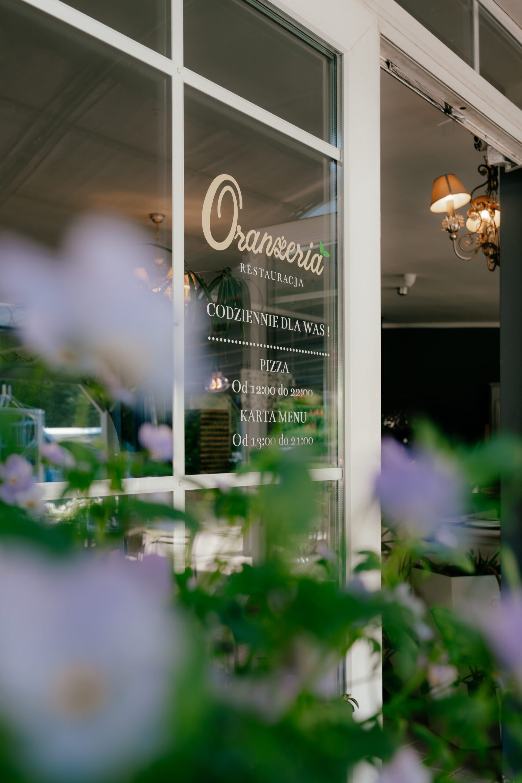 szyba restauracji Oranżeria z białym napisem i rozmytymi fioletowymi kwiatami na pierwszym planie